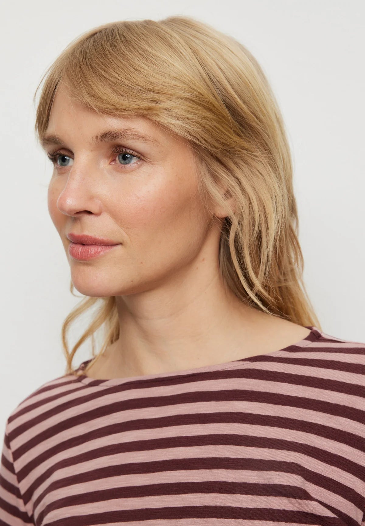 A close-up shot of a woman with blonde hair wearing a dusty rose and dark brown striped top against a light gray background.