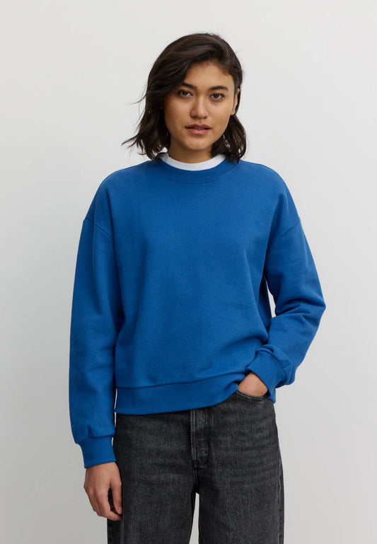 Woman wearing a blue organic cotton sweatshirt from Recolution and dark pants on a light gray background