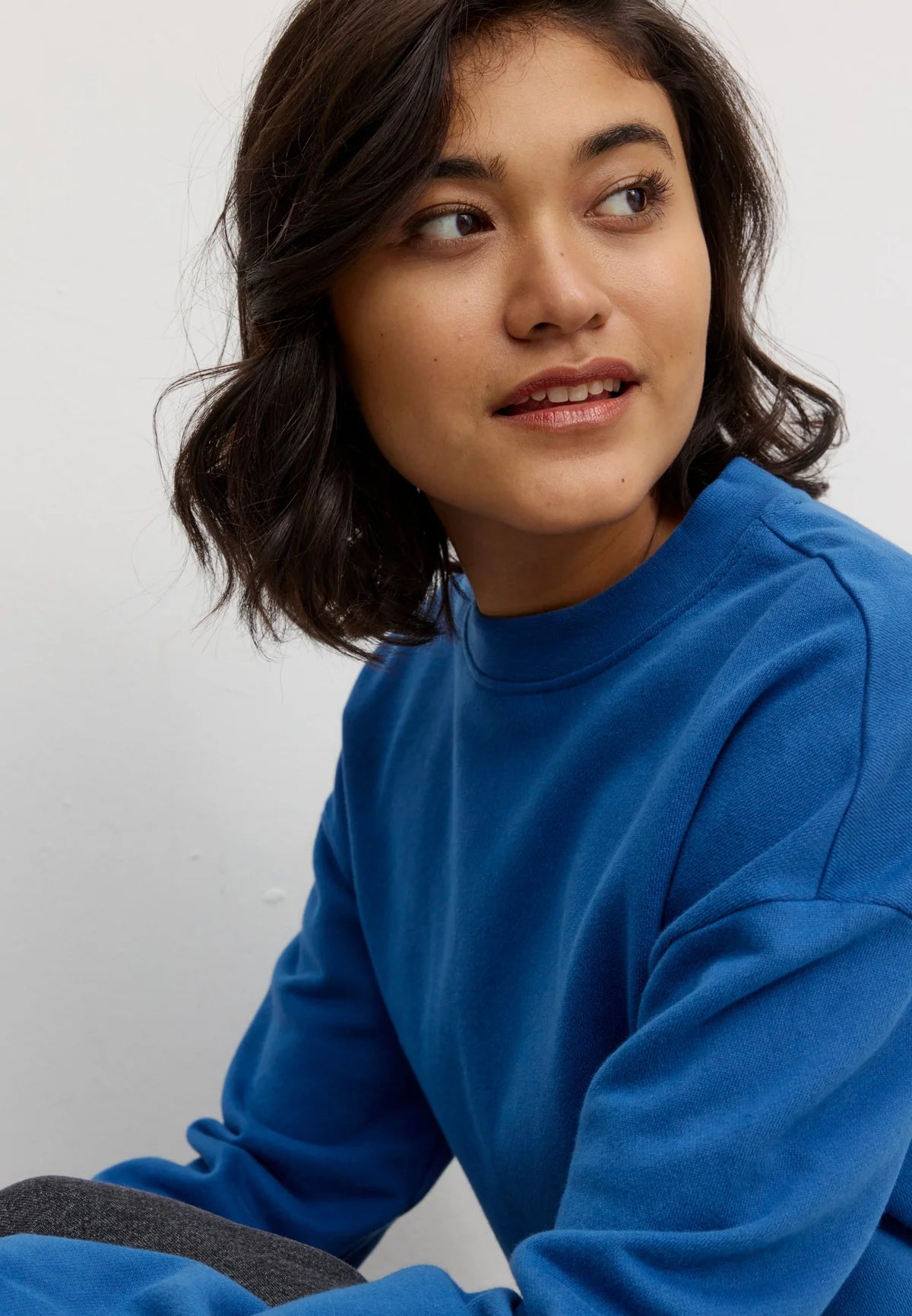 A woman with dark shoulder-length hair wearing a vibrant blue crewneck sweatshirt by Recolution, shown in a close-up profile view against a plain light gray background.