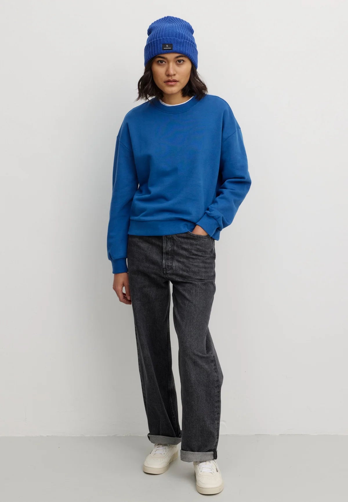 A model stands against a light gray studio background wearing a bright blue crewneck sweatshirt Recolution, a matching blue beanie, dark gray jeans, and white sneakers.