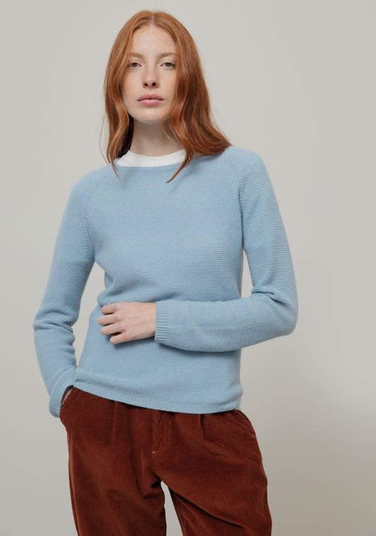 A woman with red hair wearing a light blue ribbed knit cashmere jumper from Rifo over a white crewneck shirt and brown corduroy pants, standing against a plain light gray studio background.