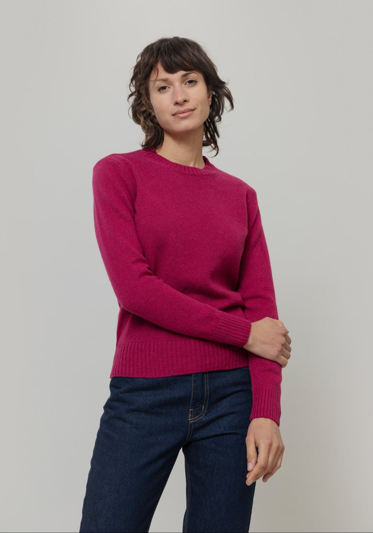 A woman modeling a magenta recycled wool crewneck knit jumper from Rifo with dark blue jeans, standing against a neutral light gray studio background.