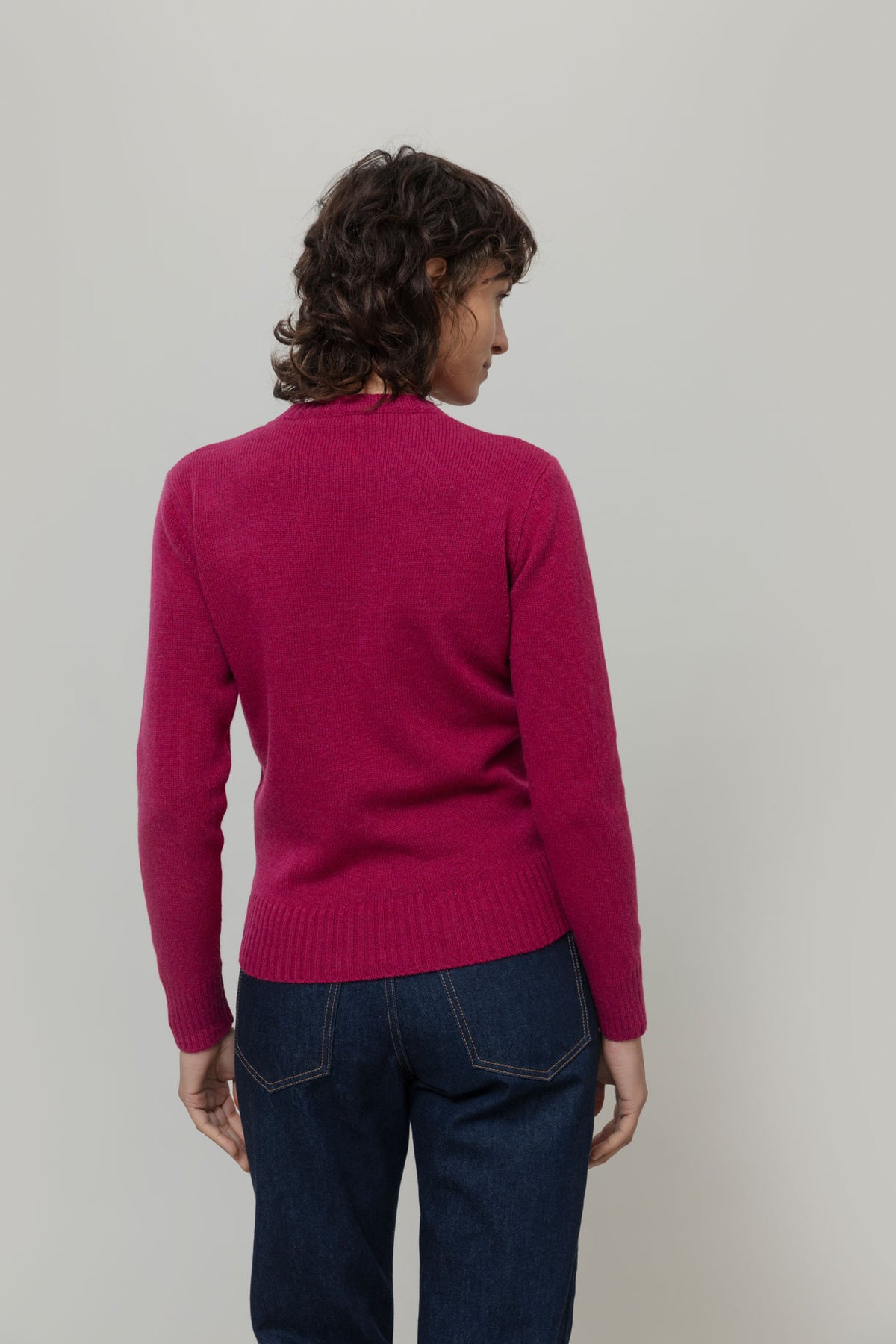 A model viewed from the back wearing a magenta pink recycled wool jumper from Rifo and dark blue jeans, standing against a plain light gray studio background.