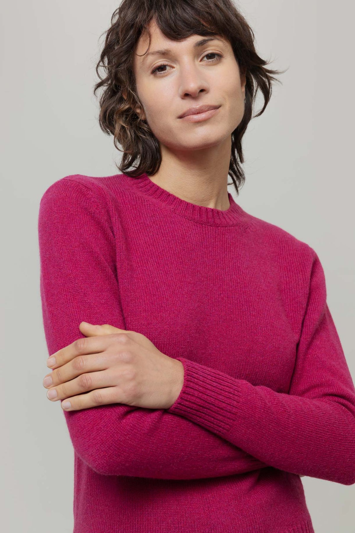 A woman with short dark hair wearing a bright magenta-pink knit crewneck jumper from Rifo, posed with her arms crossed against a solid light gray background.