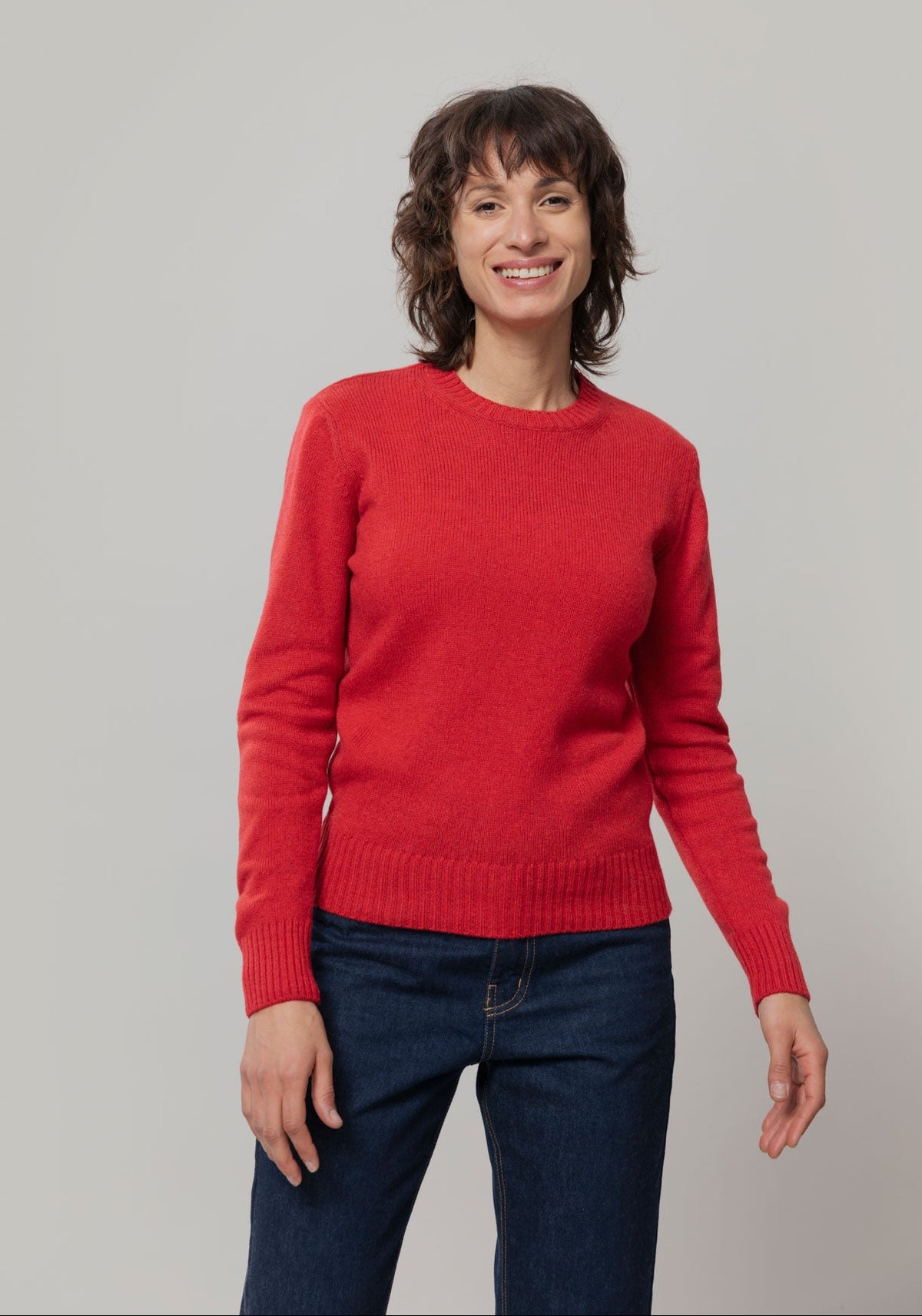 A woman wearing a bright red crewneck wool jumper by Rifo and dark blue jeans, standing against a neutral light gray studio background.