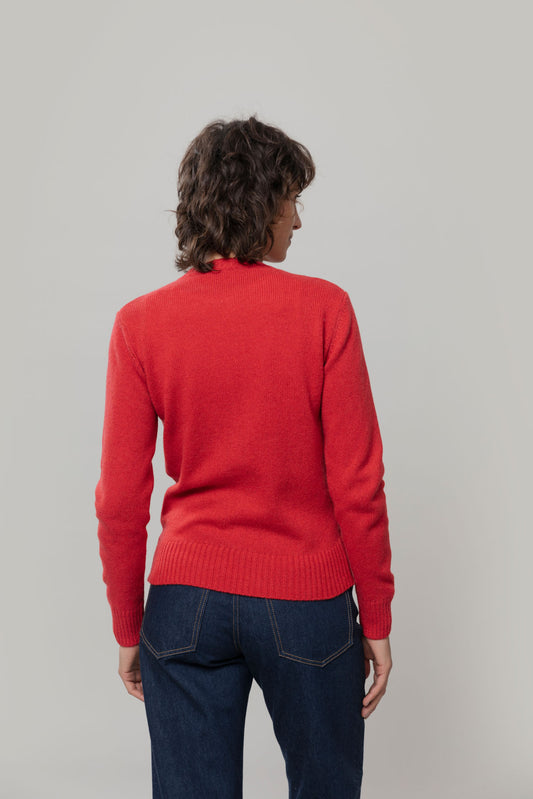 A model viewed from behind wearing a bright red wool jumper by Rifo and dark blue jeans, standing against a plain light gray studio background.