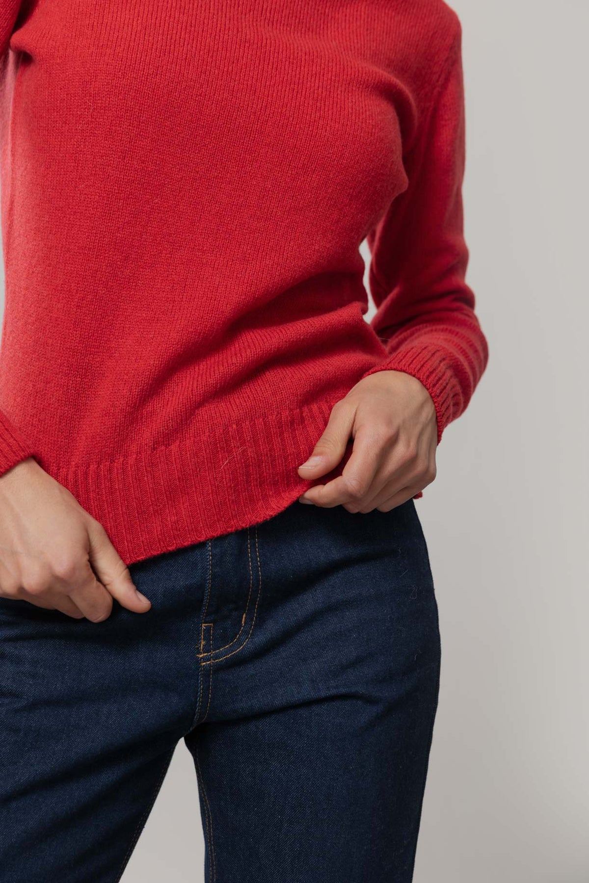 A close-up of a person wearing a bright red ribbed wool jumper and dark blue jeans, with hands pulling the hem slightly.