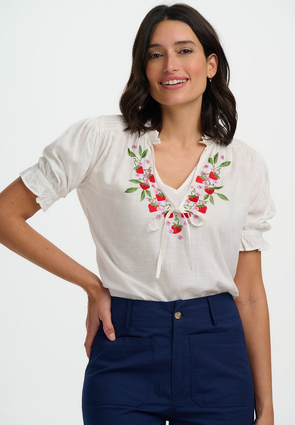 Woman wearing a white blouse with floral embroidery and navy pants on a white background