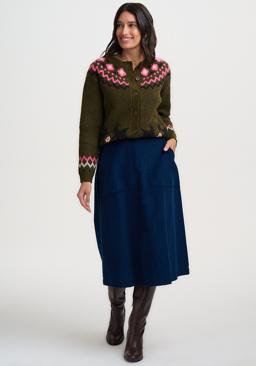 A woman modeling a dark olive green knit cardigan from Sugarhill Brighton with pink and white geometric patterns across the yoke and cuffs, paired with a dark navy midi skirt and brown knee-high boots, against a plain white studio background.