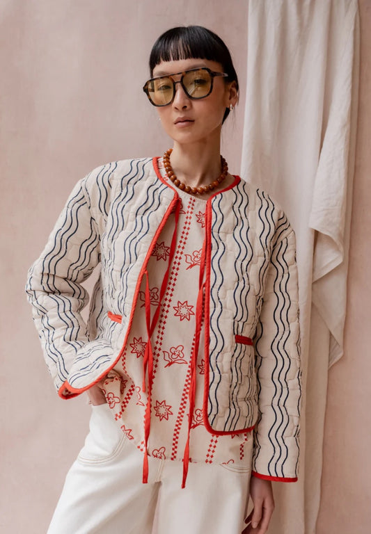 Reversible quilted jacket from TBCo in cream, with vertical wave lines in navy blue. Outer layer is made from 100% high-quality cotton, the filling is made from recycled polyester. With two front ties for adjustable styling.