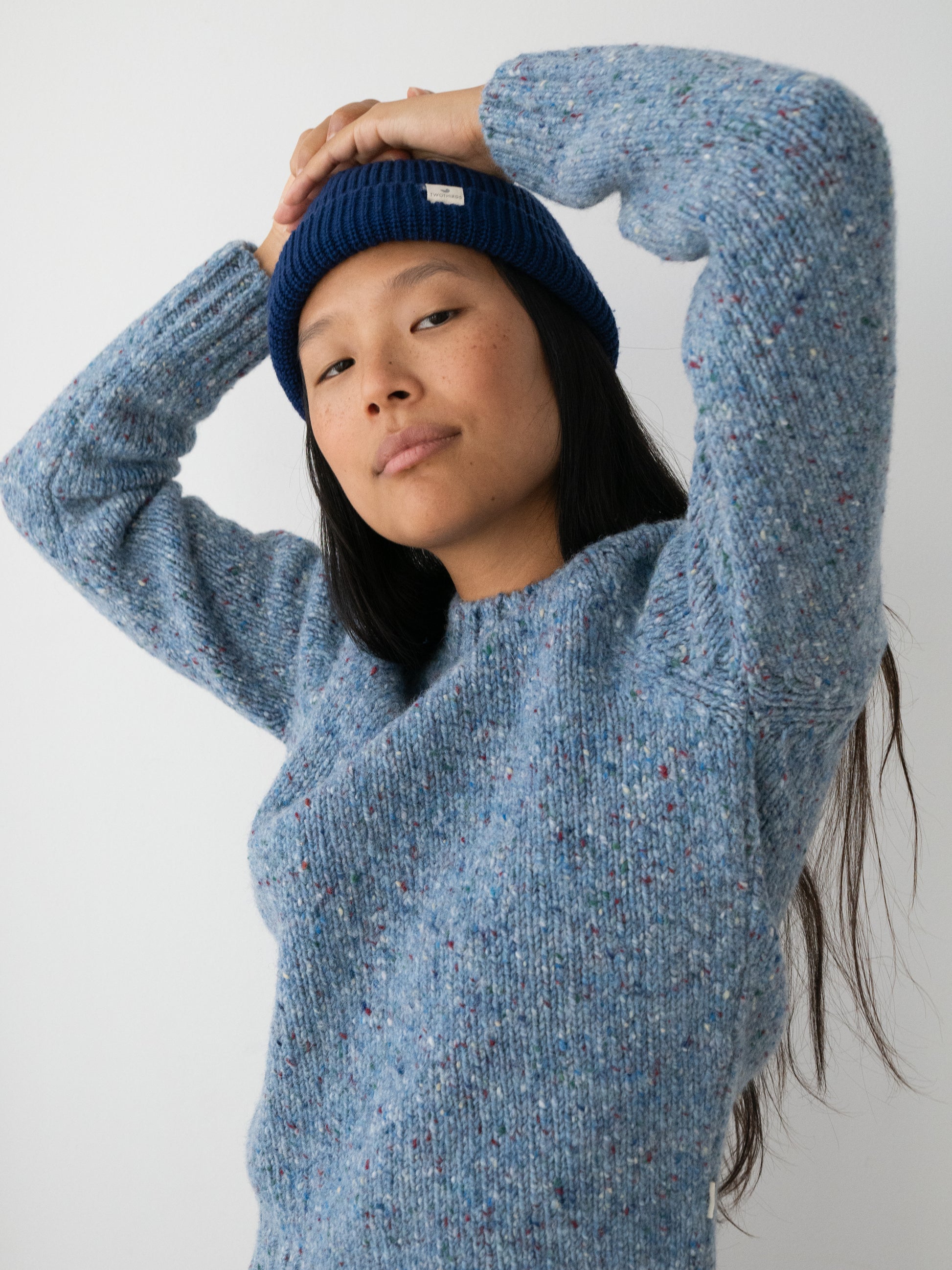 A woman wearing a textured heathered blue knit sweater by TWOTHIRDS made from Donegal yarn and a navy blue beanie, posing with her arms raised against a plain light gray background.