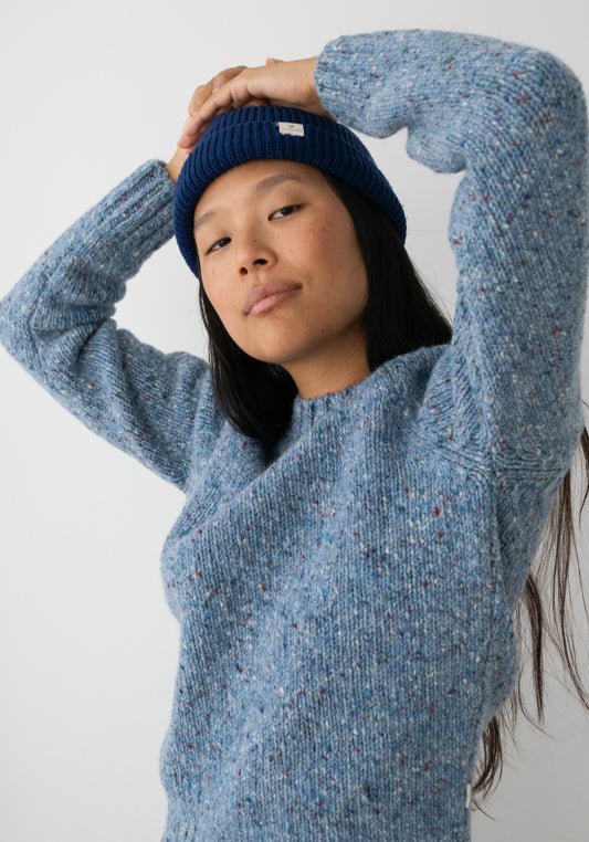 A woman wearing a textured heathered blue knit sweater by TWOTHIRDS made from Donegal yarn and a navy blue beanie, posing with her arms raised against a plain light gray background.