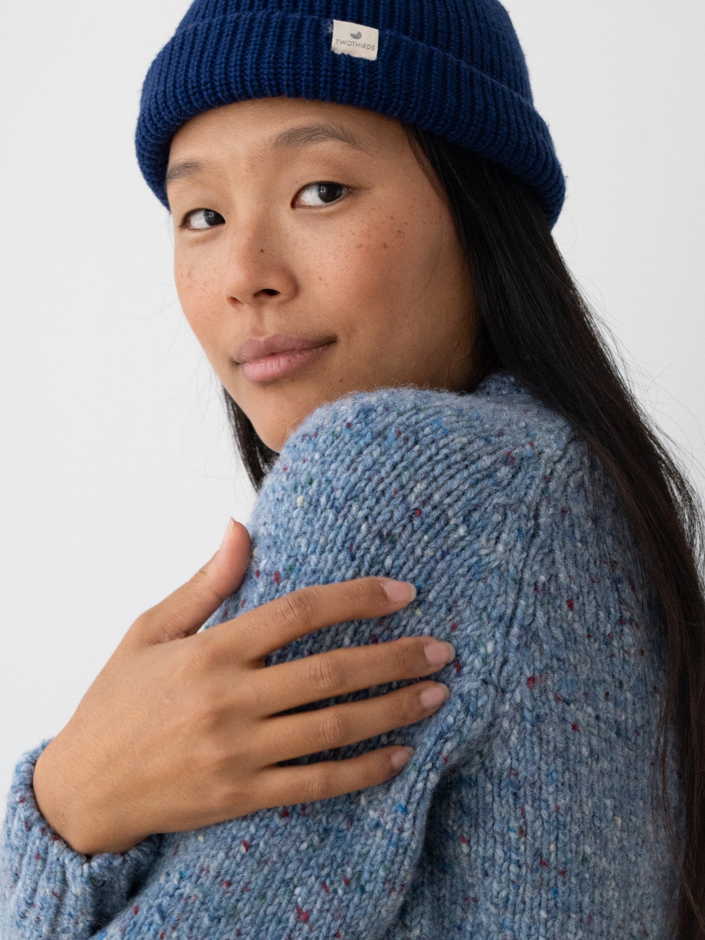 A woman wearing a navy blue ribbed knit beanie with a small white brand tag and a textured heathered blue knit sweater from TWOTHIRDS made from Donegal yarn, posing against a clean white background.