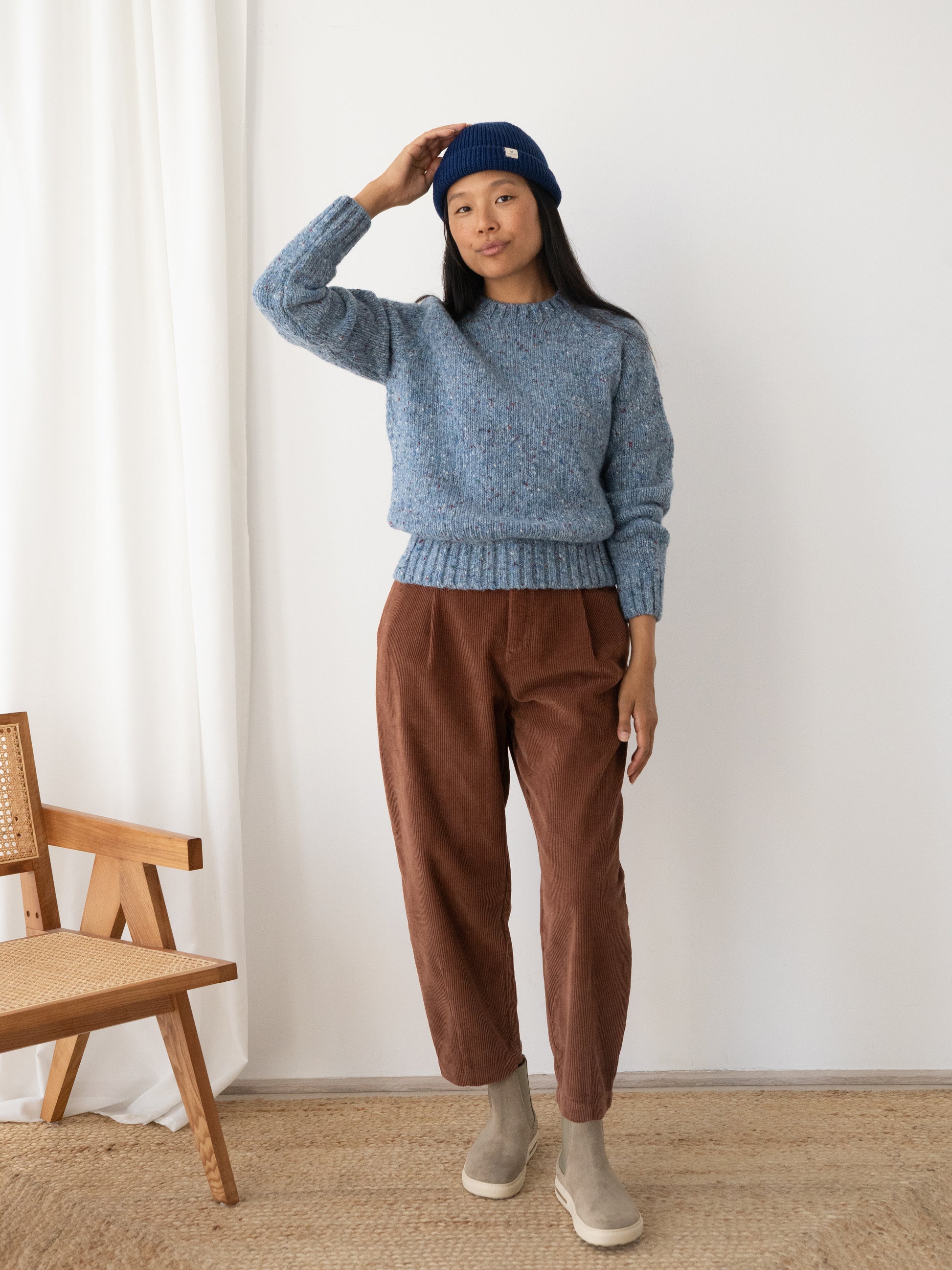 A woman stands indoors wearing a heathered blue knit sweater from TWOTHIRDS, made from Donegal wool, brown corduroy pants, gray slip-on shoes, and a navy beanie. She is posed in front of a white wall with a wooden chair partially visible on the left.