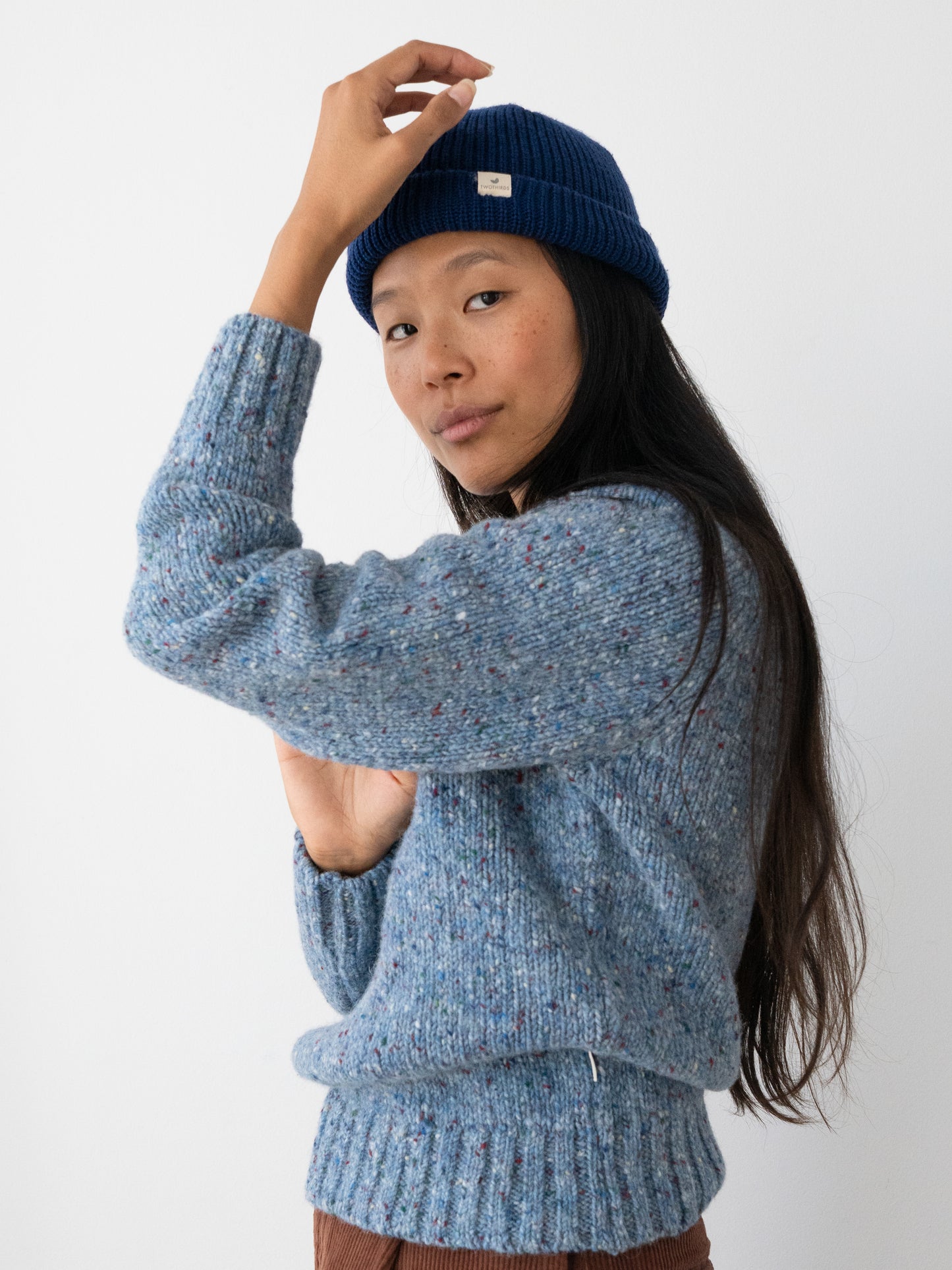 A woman wearing a blue marled knit jumper from TWOTHIRDS made from Donegal yarn and a navy blue beanie hat, posing against a plain white background.
