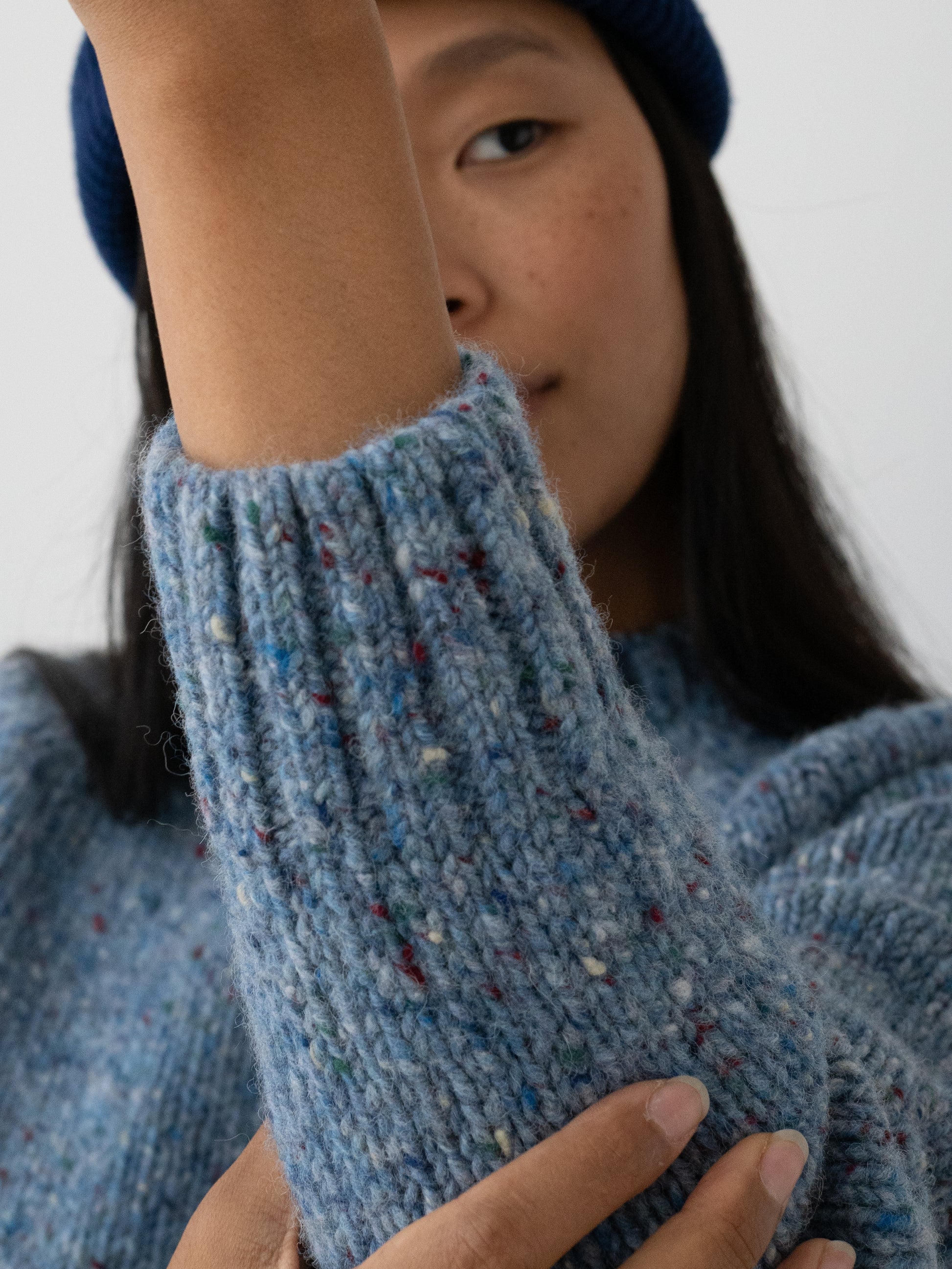 Close-up of a person wearing a chunky knit blue Donegal jumper from TWOTHIRDS with multicolored flecks, showing the ribbed cuff and textured fabric.
