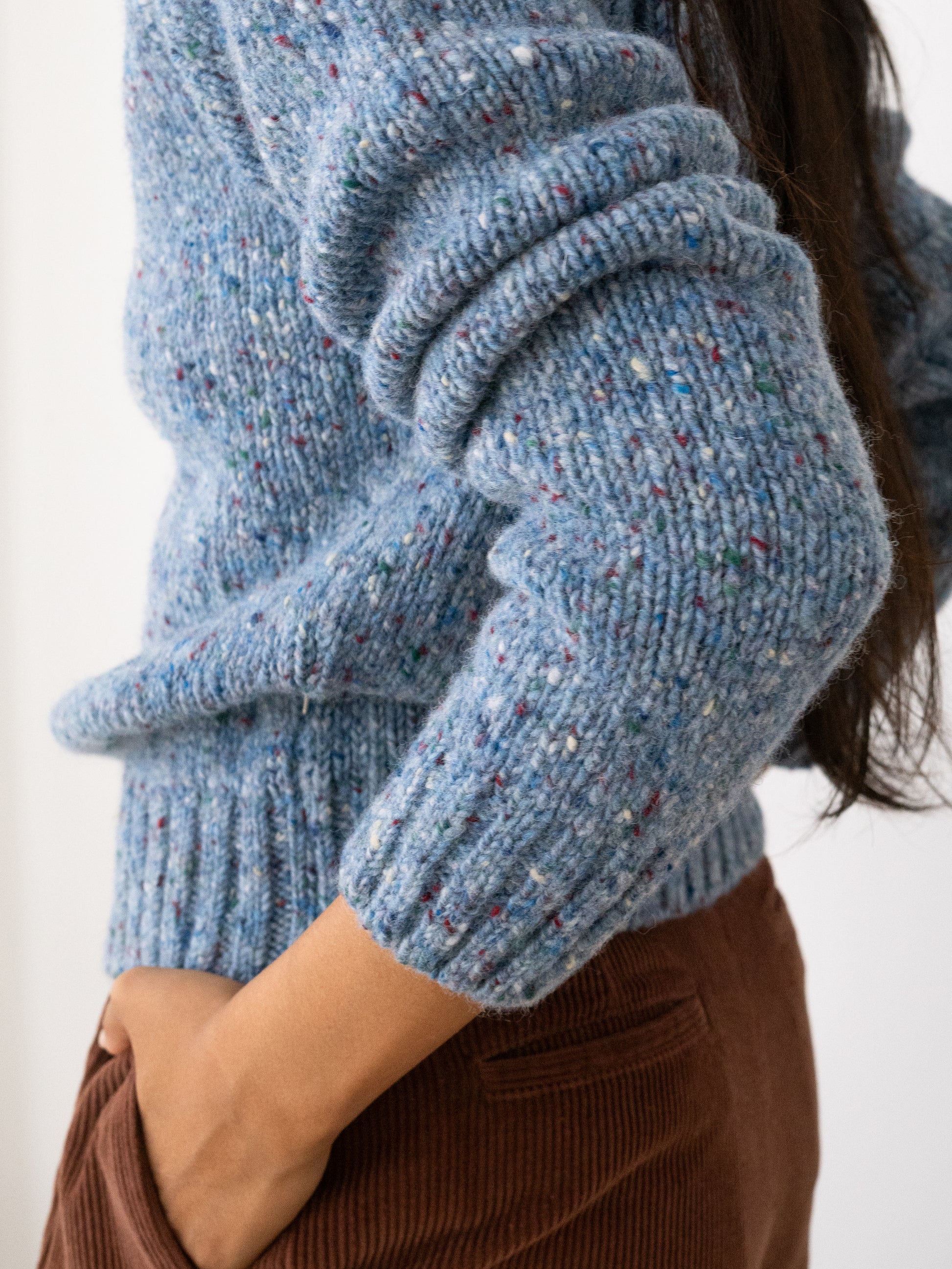 Close-up of a person wearing a chunky knit Donegal jumper from TWOTHIRDS in a heathered light blue with multicolored flecks, paired with brown corduroy pants against a plain white background.