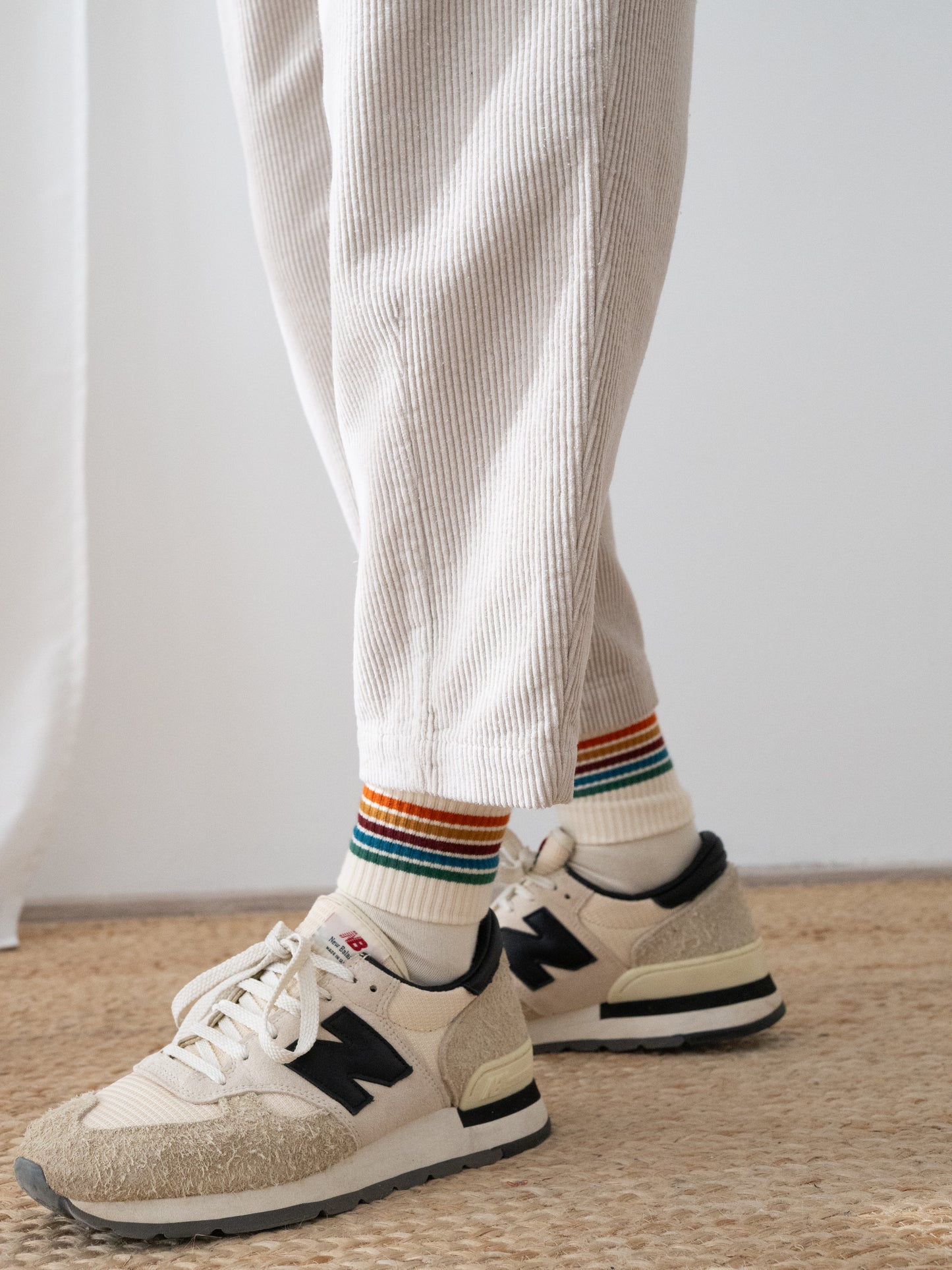 A person wearing off-white ribbed corduroy pants HAruku from TWOTHIRDS, white crew socks with rainbow stripes, and beige New Balance sneakers on a woven jute rug.