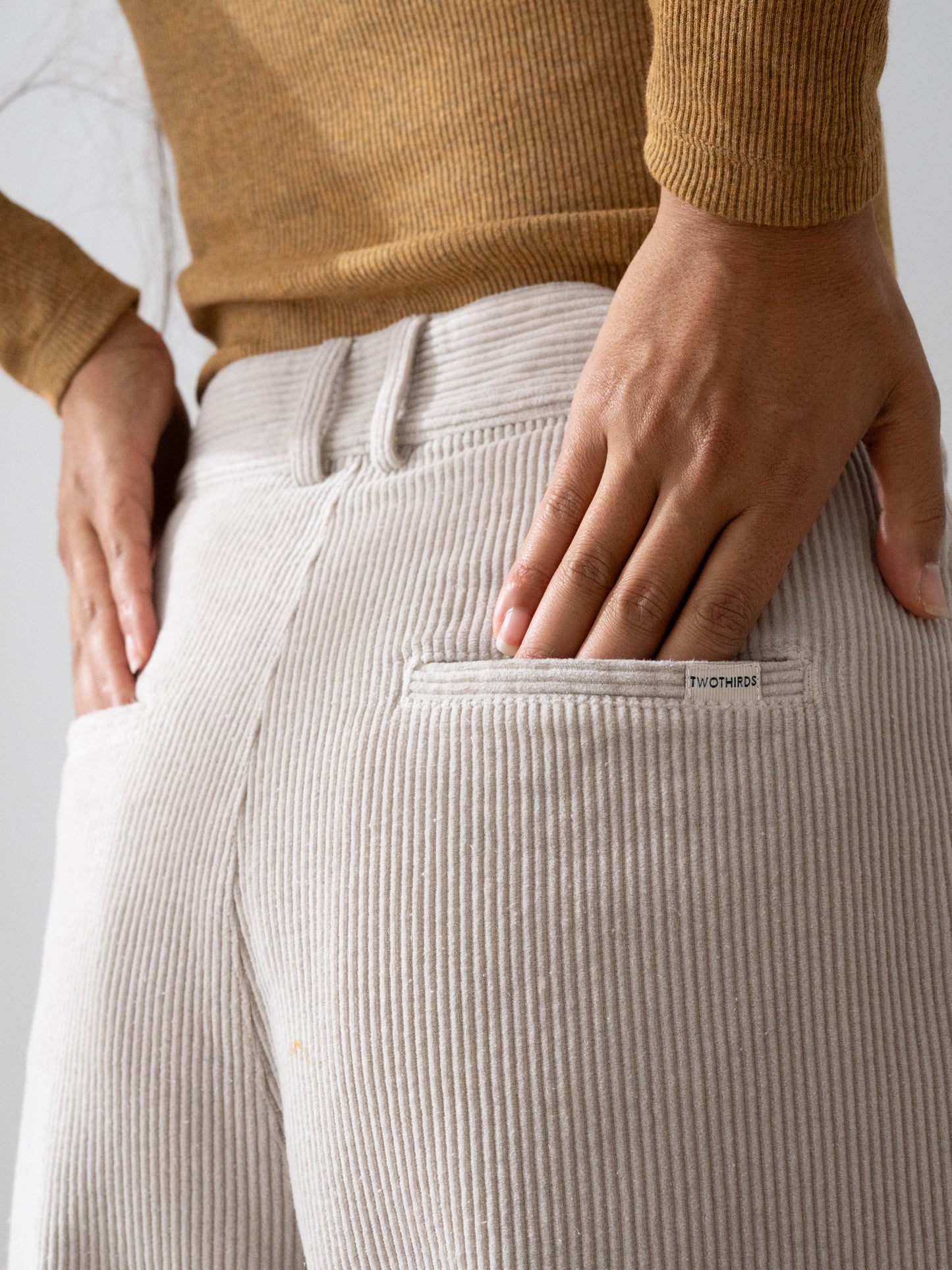 Close-up of a person wearing light beige ribbed Haruku corduroy trousers from TWOTHIRDS, showing the back pocket with a small 'fashion' label and hands resting in the pockets, paired with a tan ribbed sweater.