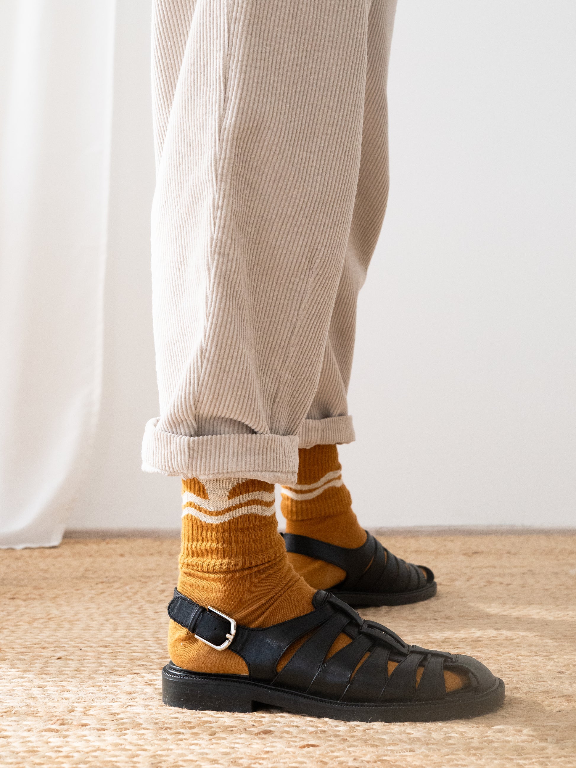A person wearing light beige Haruku corduroy pants from TWOTHIRDS, mustard yellow socks with white stripes, and black leather sandals, standing on a woven jute rug against a white curtain background.