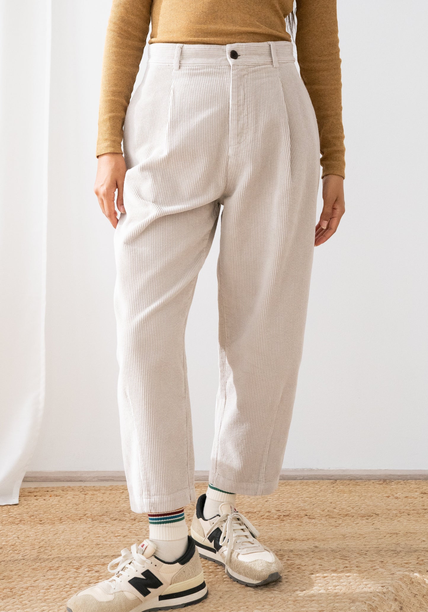 A person wearing light beige high-waisted cropped trousers, the Haruku corduroy trousers from TWOTHIRDS, a tan long-sleeve top, striped socks, and New Balance sneakers, standing indoors against a white wall and light wood floor.