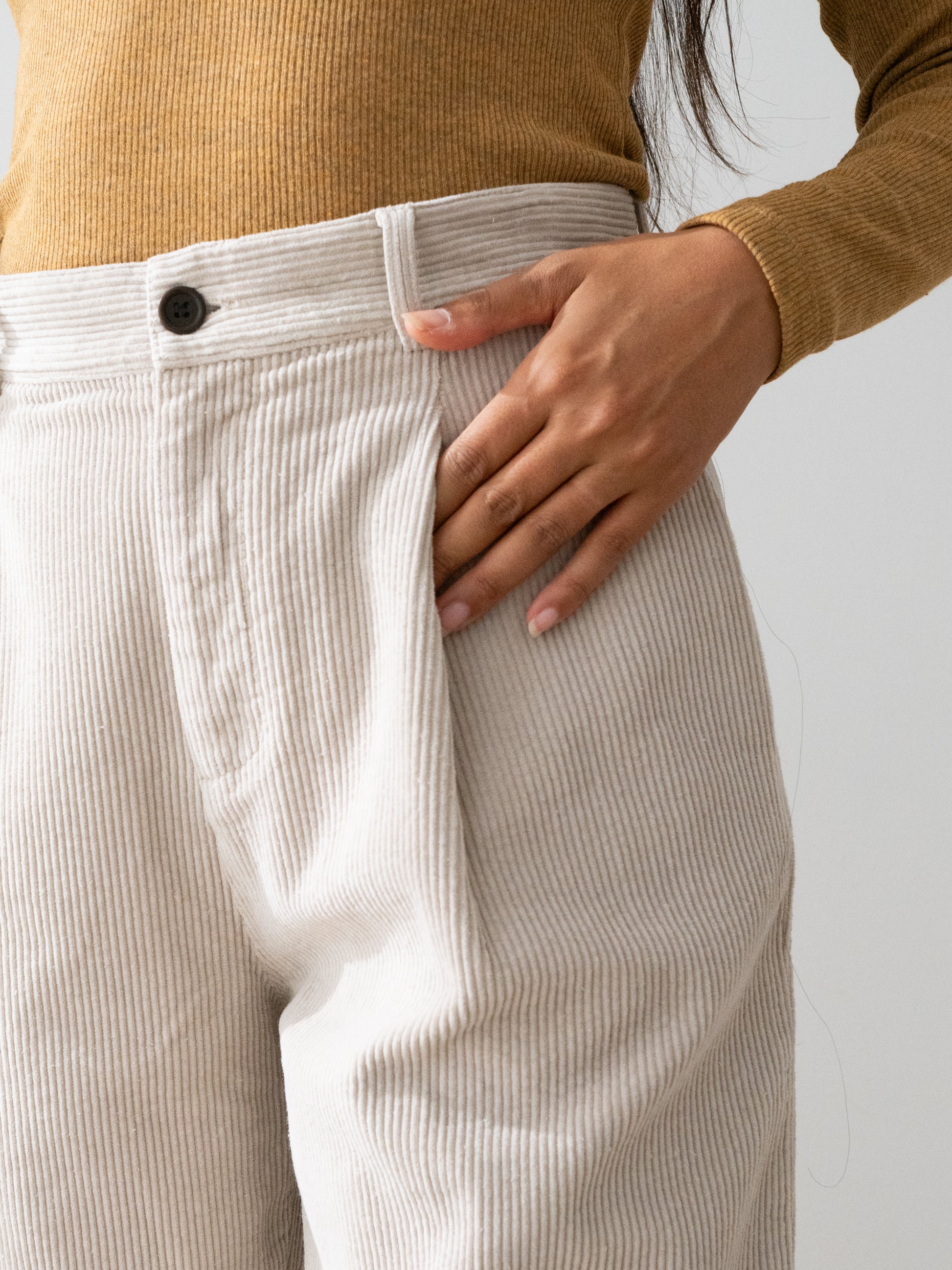 Close-up of a person wearing light beige Haruku corduroy trousers from TWOTHIRDS and a mustard yellow ribbed sweater, with one hand resting on the hip pocket.