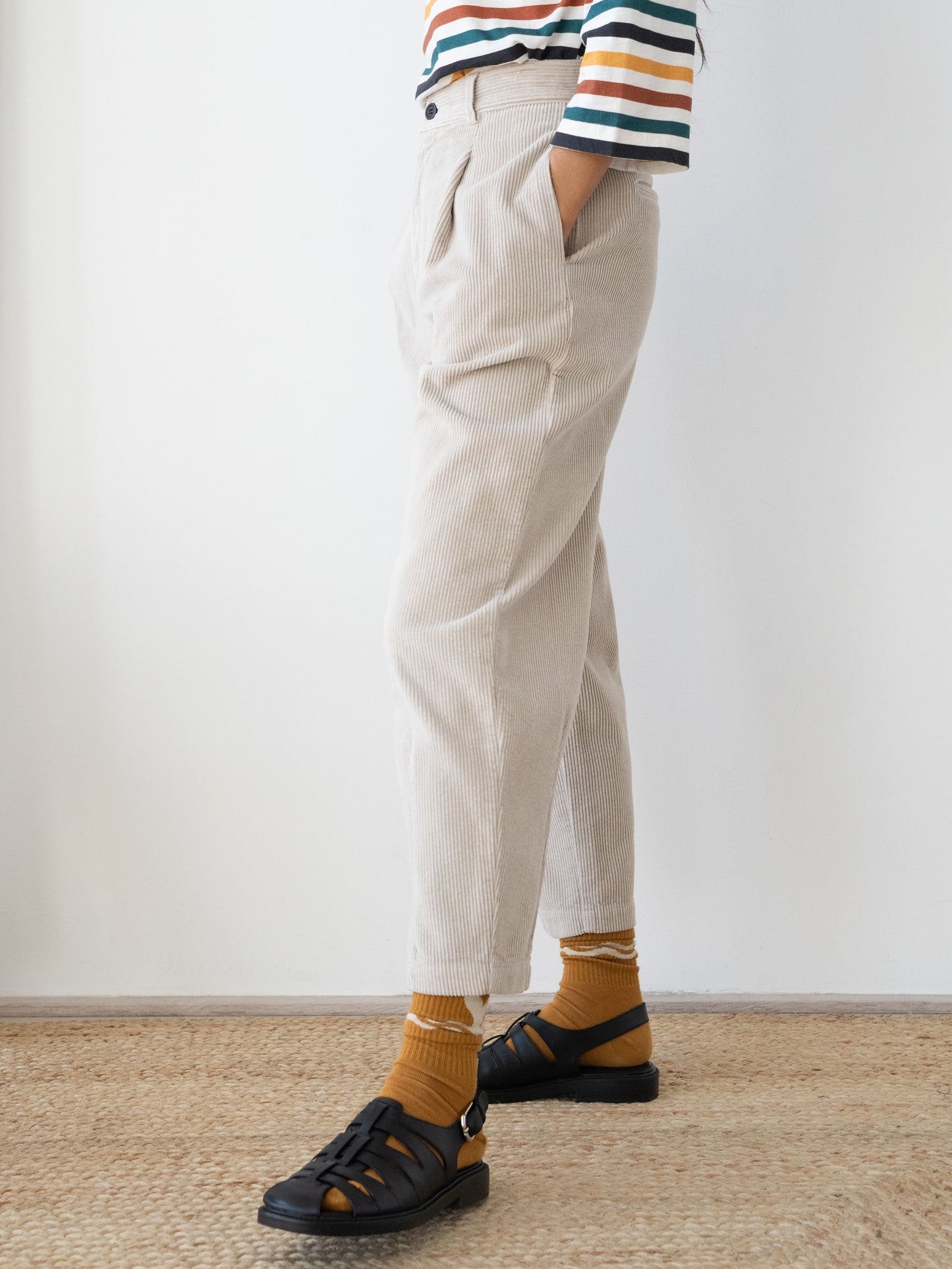 A person wearing light beige  Haruku corduroy trousers from TWOTHIRDS, mustard yellow socks, and black sandals, standing on a woven jute rug against a plain white wall.