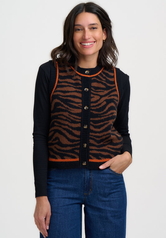 A woman modeling a sleeveless brown and black tiger-striped knit vest by Sugarhill Brighton over a black long-sleeve top, paired with blue jeans, against a plain light grey background.