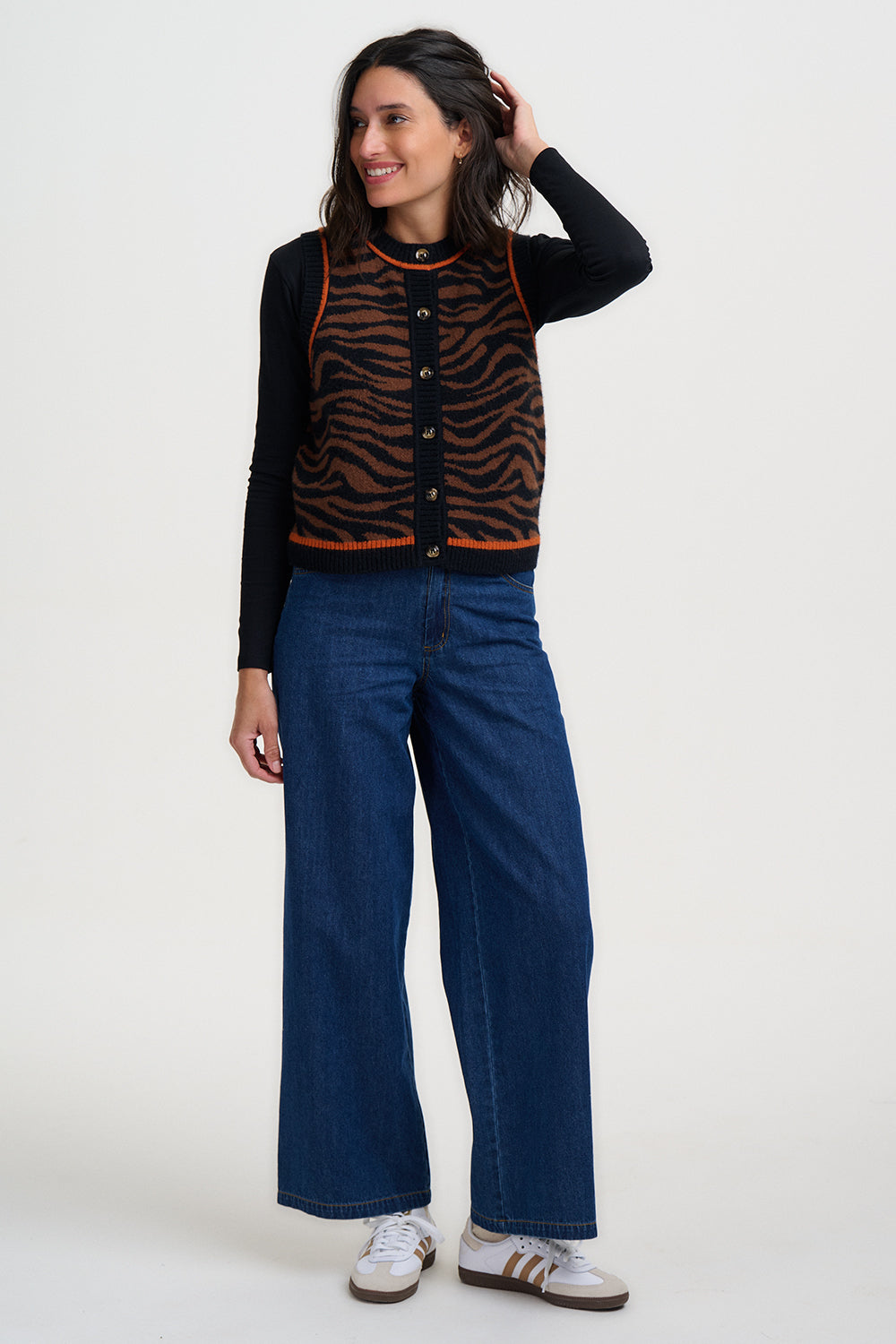 Woman wearing a black and brown knitted gilet from Sugarhill Brighton with orange piping and blue jeans on a white background